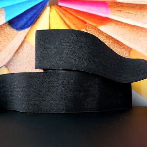 Schwarzes, 5cm breites Gummiband mit eingewebtem Jacquard-Blumenmuster als aufgewickeltes Band vor buntem Stifthintergrund präsentiert auf Spitzentraum.ch.