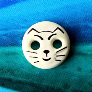 Runder, 12mm grosser Motivknopf in ecrufarbener Butterfarbe mit aufgedrucktem schwarzem Katzengesicht und zwei L&ouml;chern als Augen vor blauem Studiohintergrund.