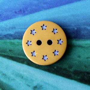 Runder, ockerfarbener Kunststoffknopf (18mm) mit kreisf&ouml;rmig angeordneten, kleinen blauen Sternen auf blau-gr&uuml;nem Hintergrund bei Spitzentraum.ch.