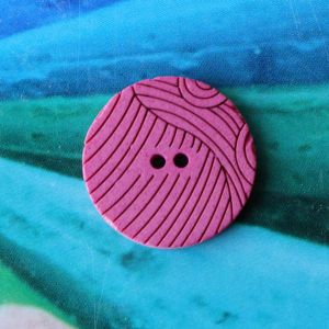 Runder, flacher Kunststoffknopf (18mm) in Pink mit grafischem, geschwungenem Streifenmuster auf blau-gr&uuml;nem Hintergrund bei Spitzentraum.ch.