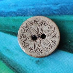 Einzelner 17mm Kokosnuss-Knopf mit dunkel nachgedunkeltem Mandala-Muster auf der hellen Vorderseite pr&auml;sentiert vor blau-gr&uuml;nem Grund bei Spitzentraum.ch.